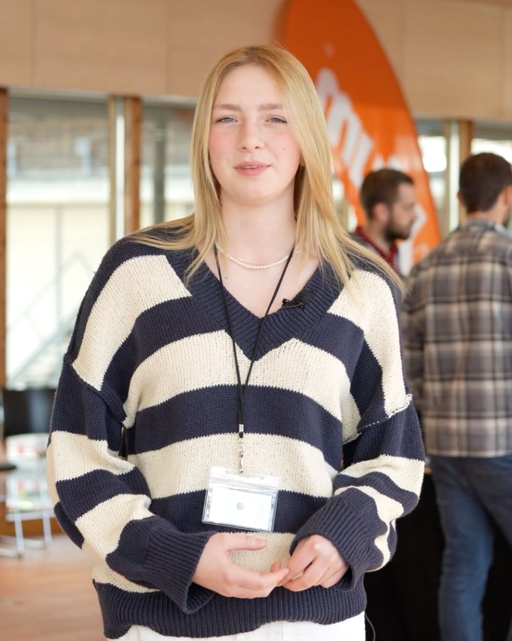 Junge blonde Frau mit Namensschild steht vor einem orangefarbenen Banner