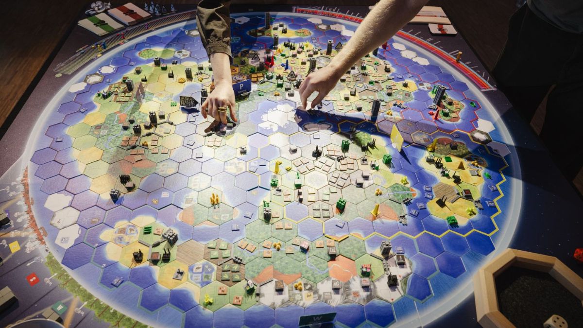 Vogelperspektive auf ein rundes Spielbrett mit Hexfeldern, Spielfiguren und Karten