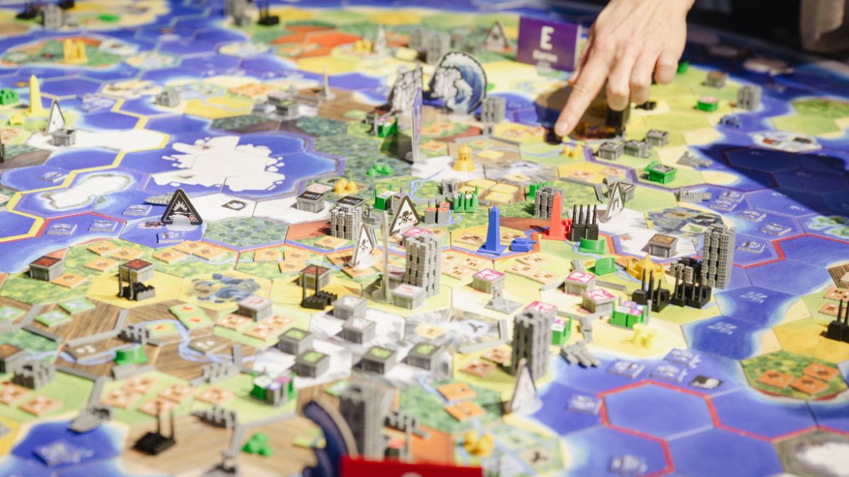 Vogelperspektive auf ein Spielbrett mit Küsten, Städten und Klimaelementen, eine Hand zeigt auf das Brettf