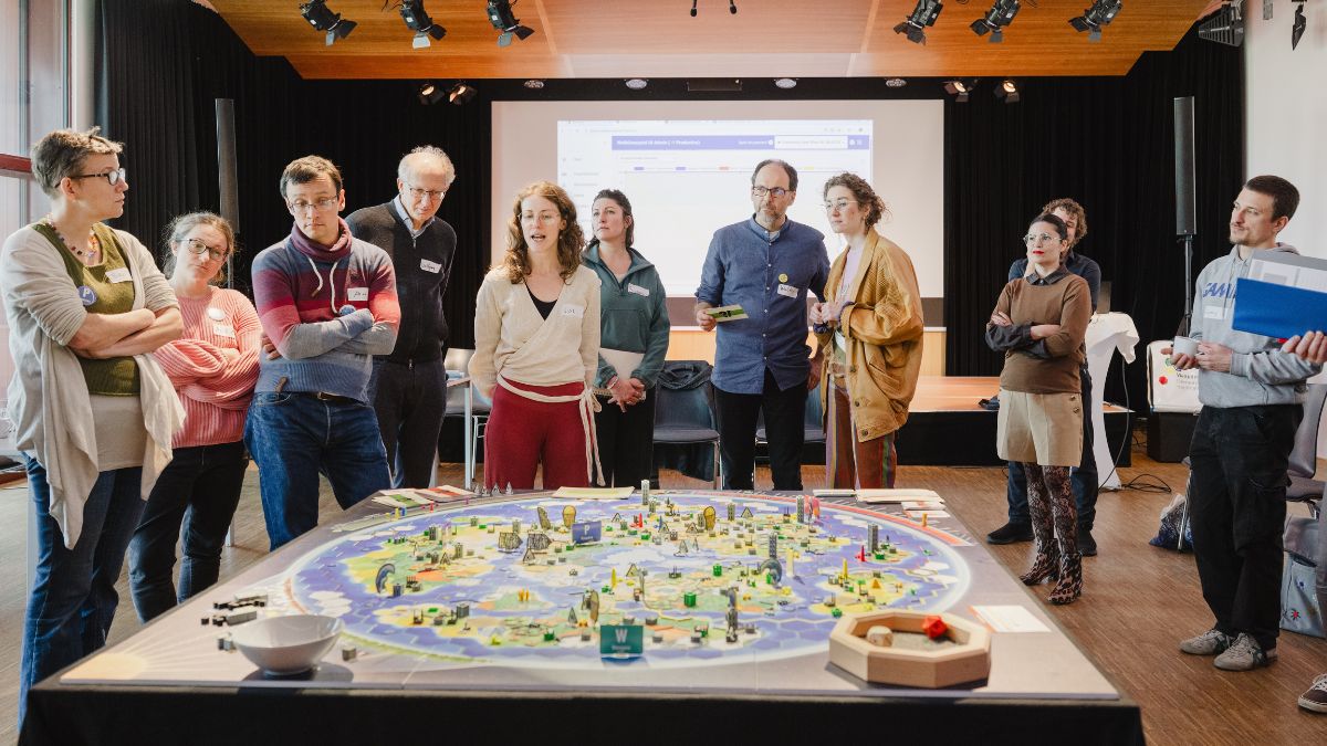 Gruppe von Personen steht vor einem Spielbrett in einem Seminarraum, im Hintergrund eine Präsentationsleinwand