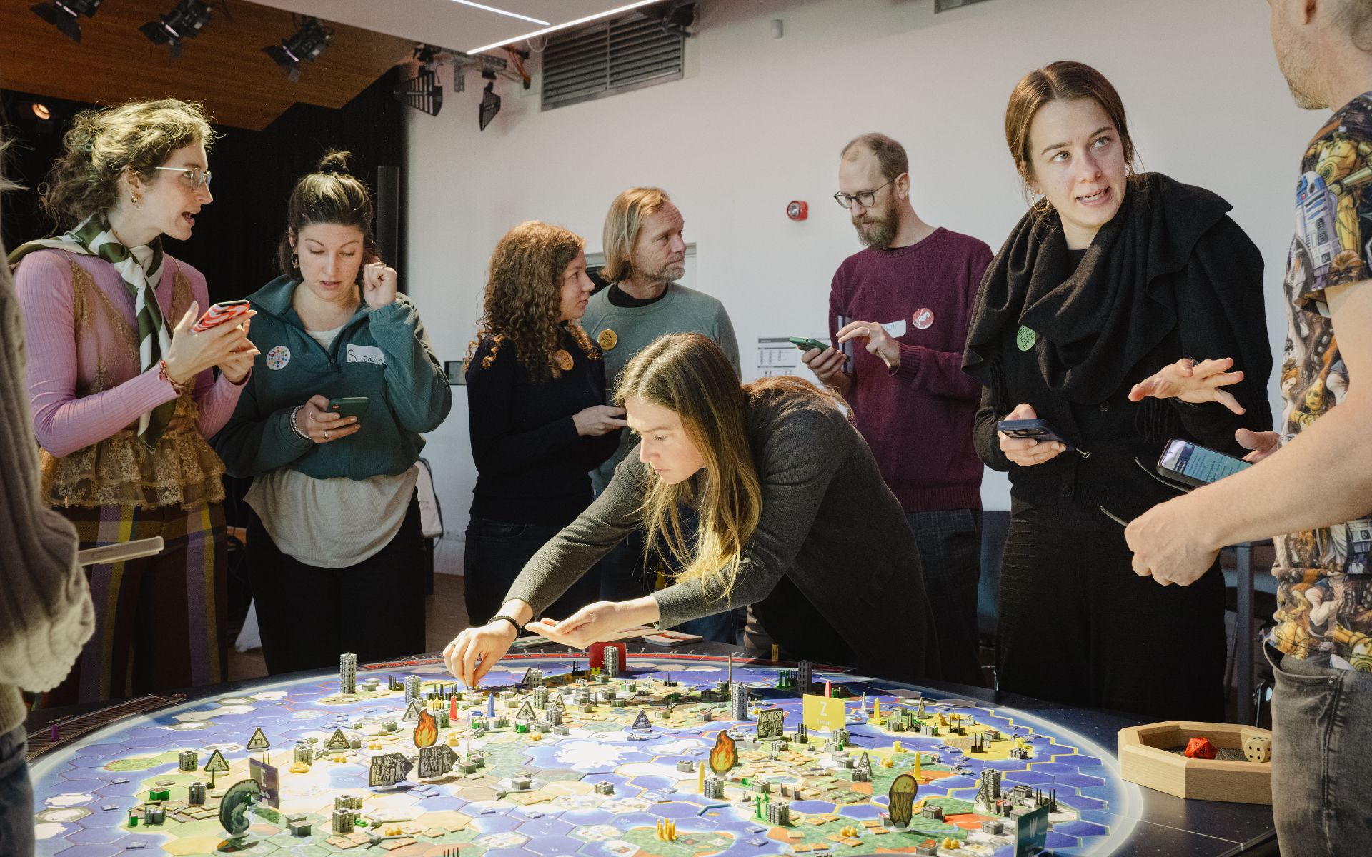 Mehrere Personen stehen um einen Tisch und spielen ein Brettspiel mit Weltkarte, Figuren und Karten