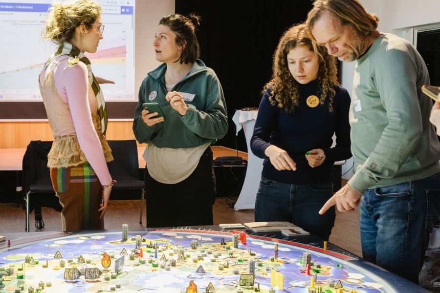 Gruppe von vier Personen steht um das beleuchtete Weltklimaspiel-Spielbrett in einem Seminarraum
