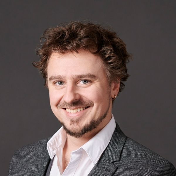 Mann mit braunen Haaren und Bart l&auml;chelt vor grauem Hintergrund